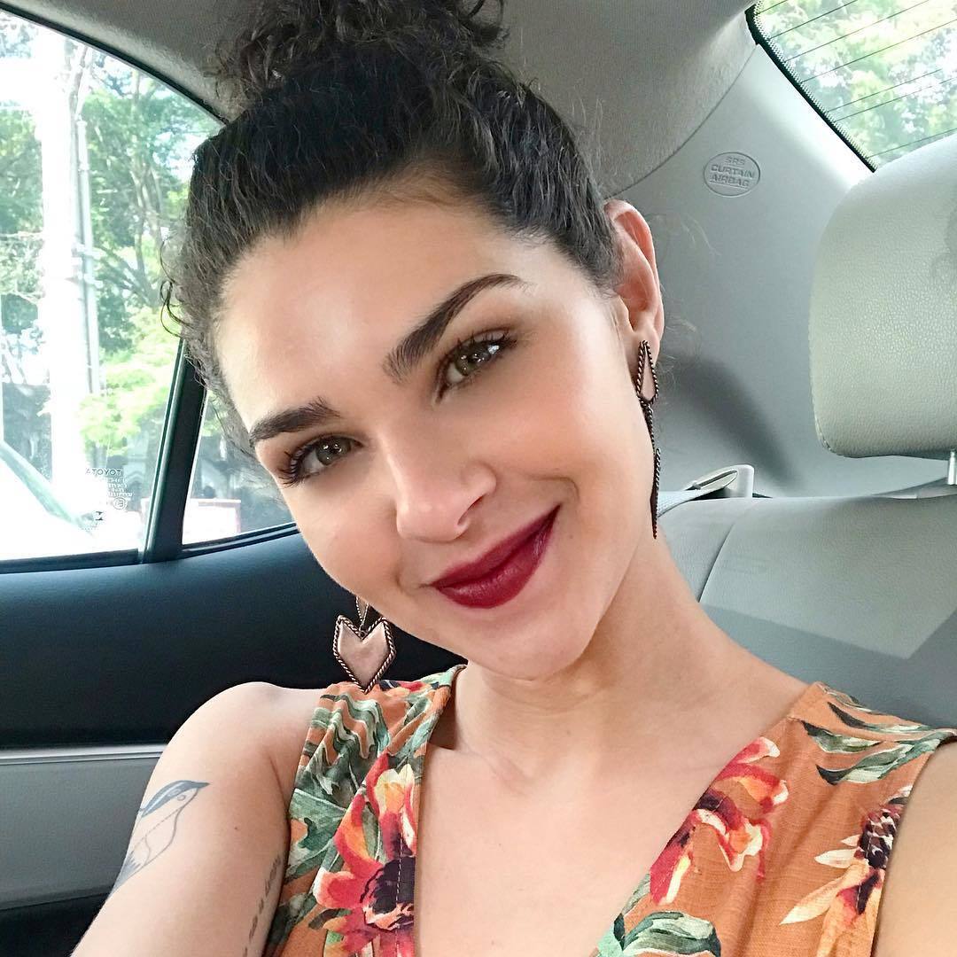A influenciadora Gabi Vasconcellos com o cabelo cacheado preso em um coque, batom vinho e top floral