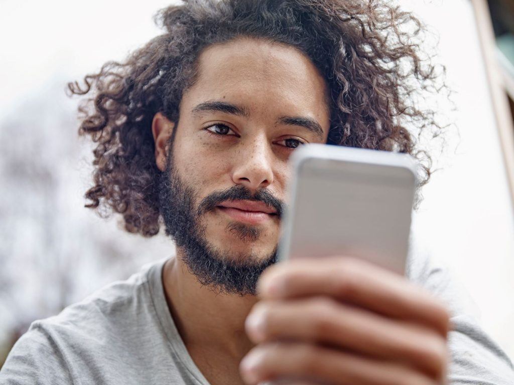 Homem de cabelos cacheados de comprimento médio olha para o celular, em suas mãos