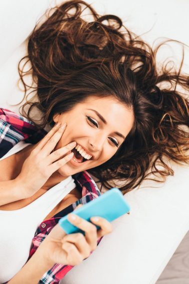Mulher deitada no sofá segura o celular na mão e sorri