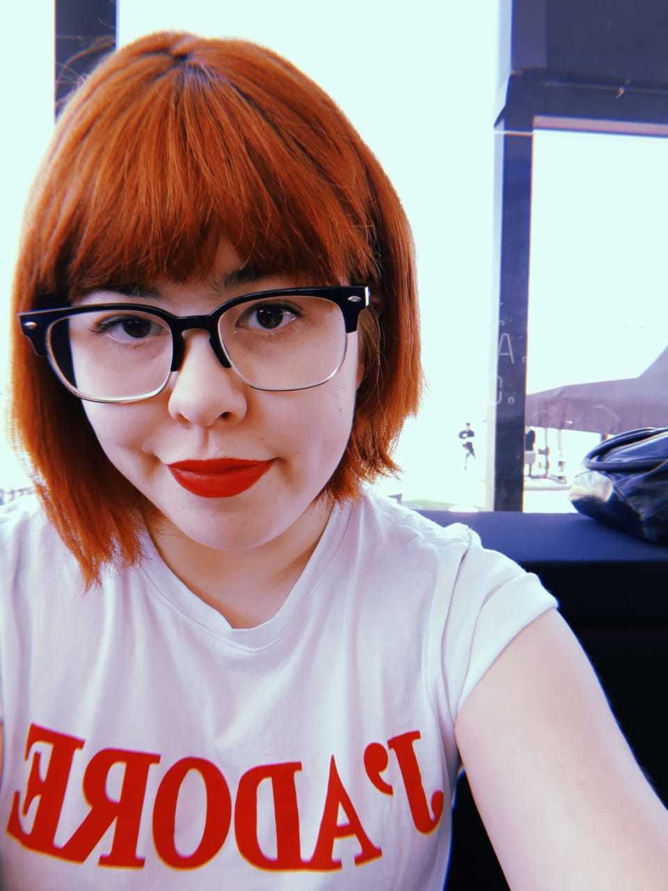 Mulher com cabelo curto com franja ruivo com óculos de grau, camiseta branca e batom vermelho