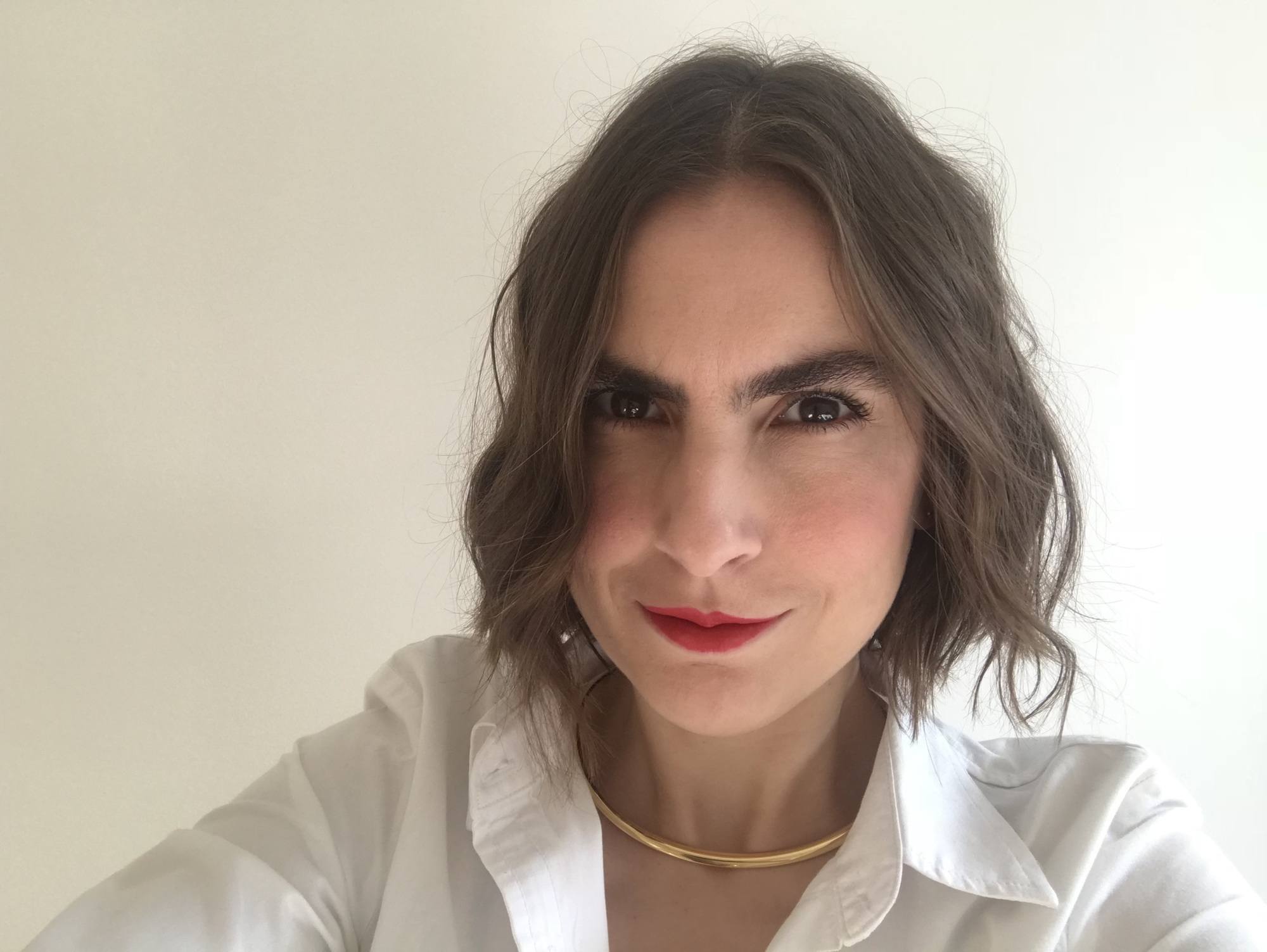 Mulher com cabelo curto castanho ondulado, batom vermelho, camisa branca e colar dourado