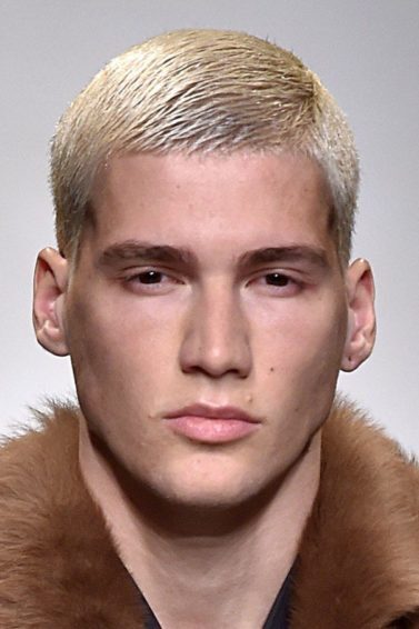 Modelo loiro desfila usando colete de pele marrom, camisa azul escura e usa cabelo masculino sombreado