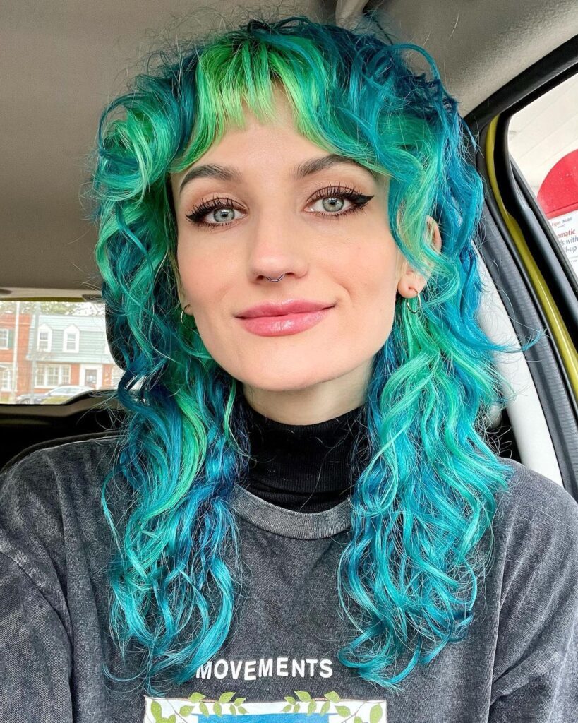Mulher de camiseta cinza dentro do carro com mullet feminino longo no cabelo colorido azul e verde estilo wolf cut com franja