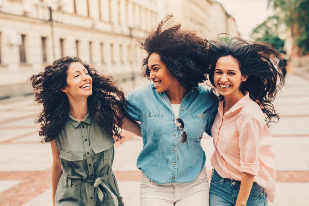 Mulheres com cabelo liso, cacheado e crespo se abraçam e caminham juntas