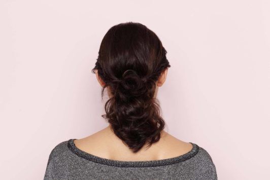Mulher de cabelo preto e rabo de cavalo usa blusa cinza e posa de costas