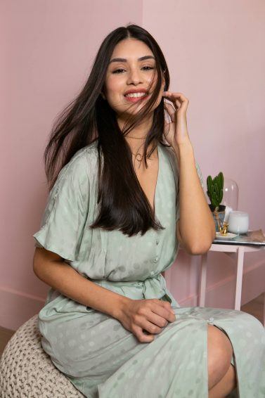 modelo de cabelo liso e vestido verde sorri para a câmera com cabelos esvoaçantes. ela usa vestido verde e está numa slaa rosa