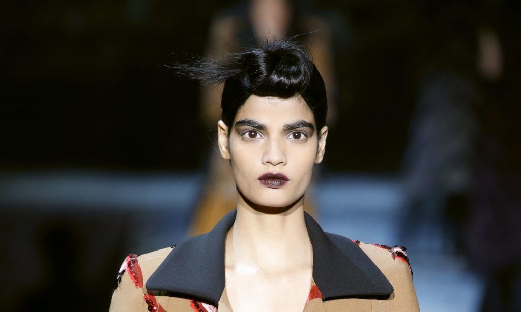 Mulher usa cabelo preto preso num coque, batom escuro e casaco cáqui, preto e vermelho.