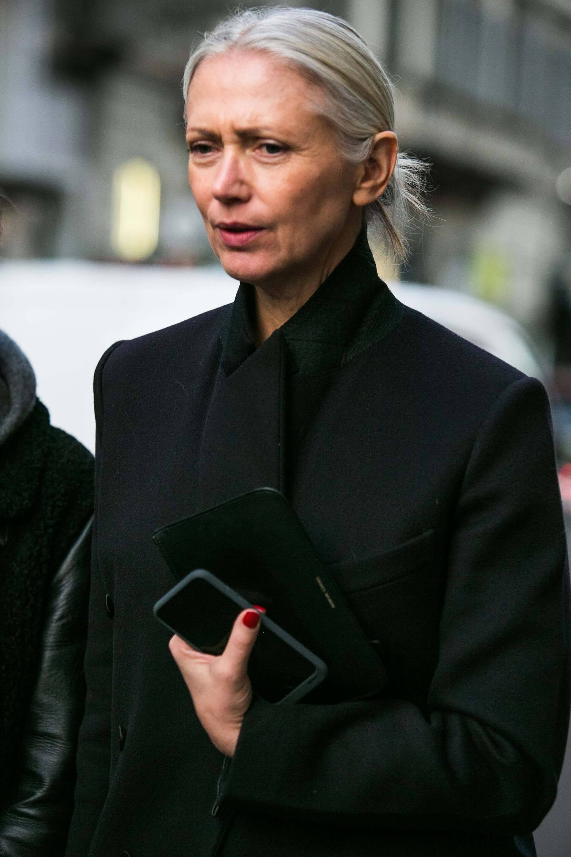 Mulher com cabelo grisalho usa cabelo preso com um nó e roupa toda preta, com casaco também preto