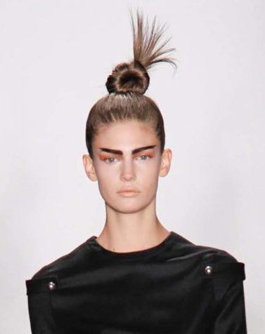 Mlulher usa o cabelo preso com um nó num coque alto e roupa toda preta, na passarela