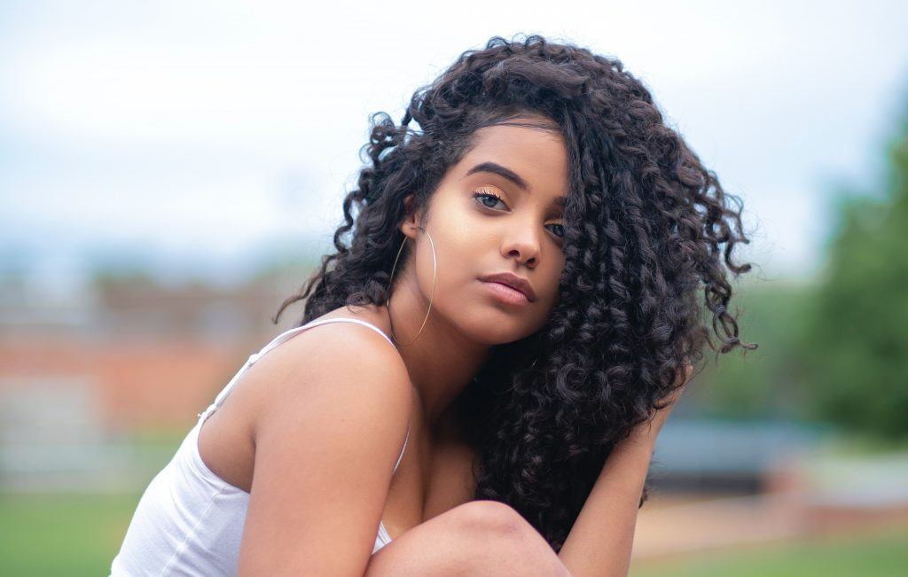 Garota jovem de cabelos cacheados longos está sentada na grama, ao ar livre. Ela está com uma das mãos no cabelo e olhando para a câmera