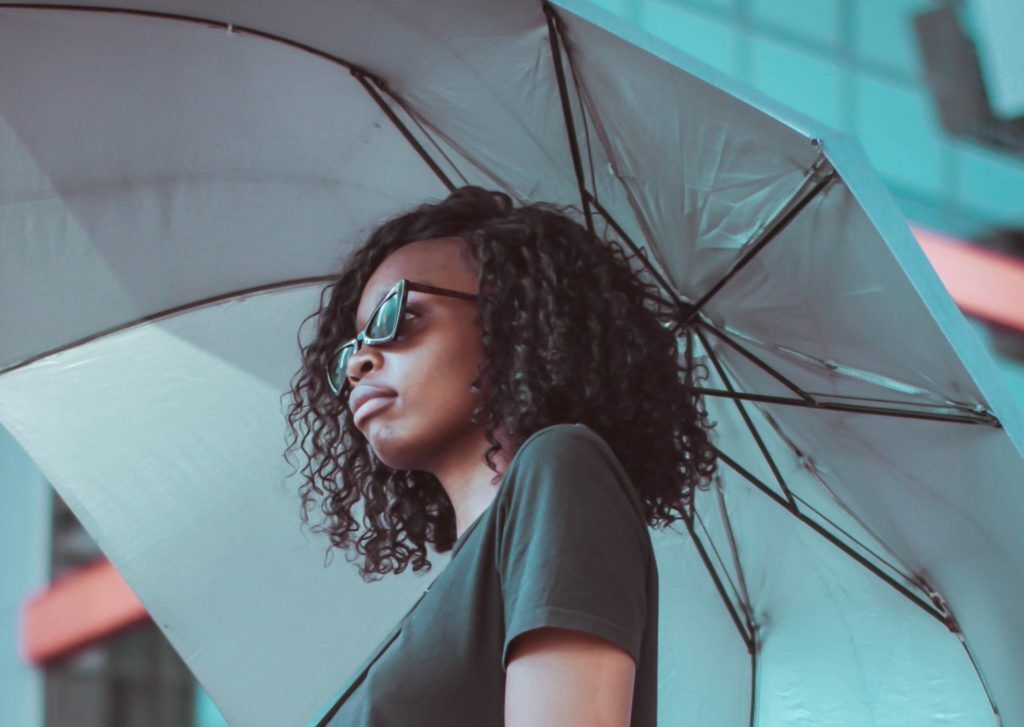 Mulher de cabelo curto cacheado se protege do sol com guarda-chuva. Ela é vista de lado e usa óculos escuros