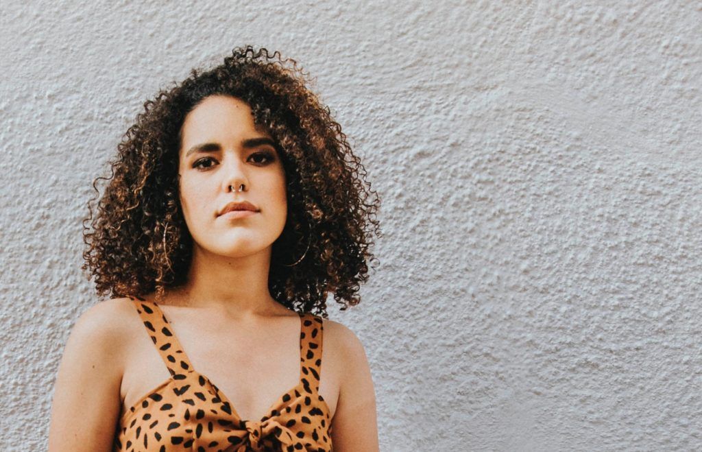 Mulher de cabelo cacheado e pele branca olhando para câmera. Ela está em frente a uma parede e usa piercing no nariz