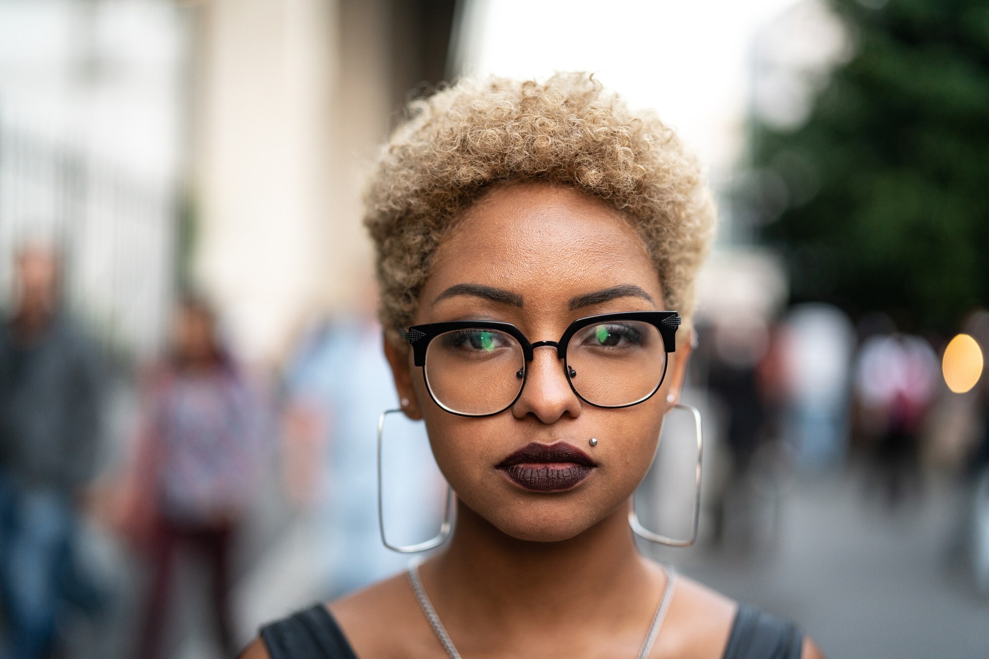 Mulher posa ao ar livre com cabelo curto big chop cacheado loiro enquanto usa óculos