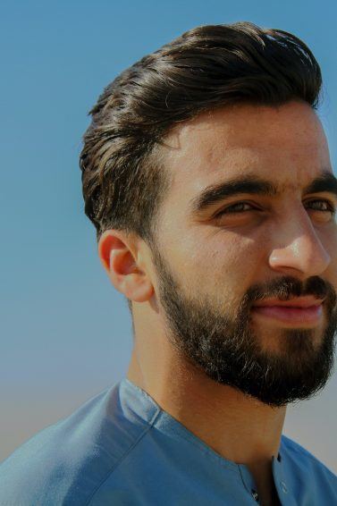Homem de barba e cabelo pretos e camisa azul, ao ar livre