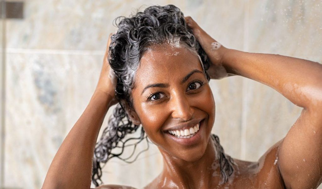 Mulher sorri com espuma de shampoo nos cabelos durante o banho