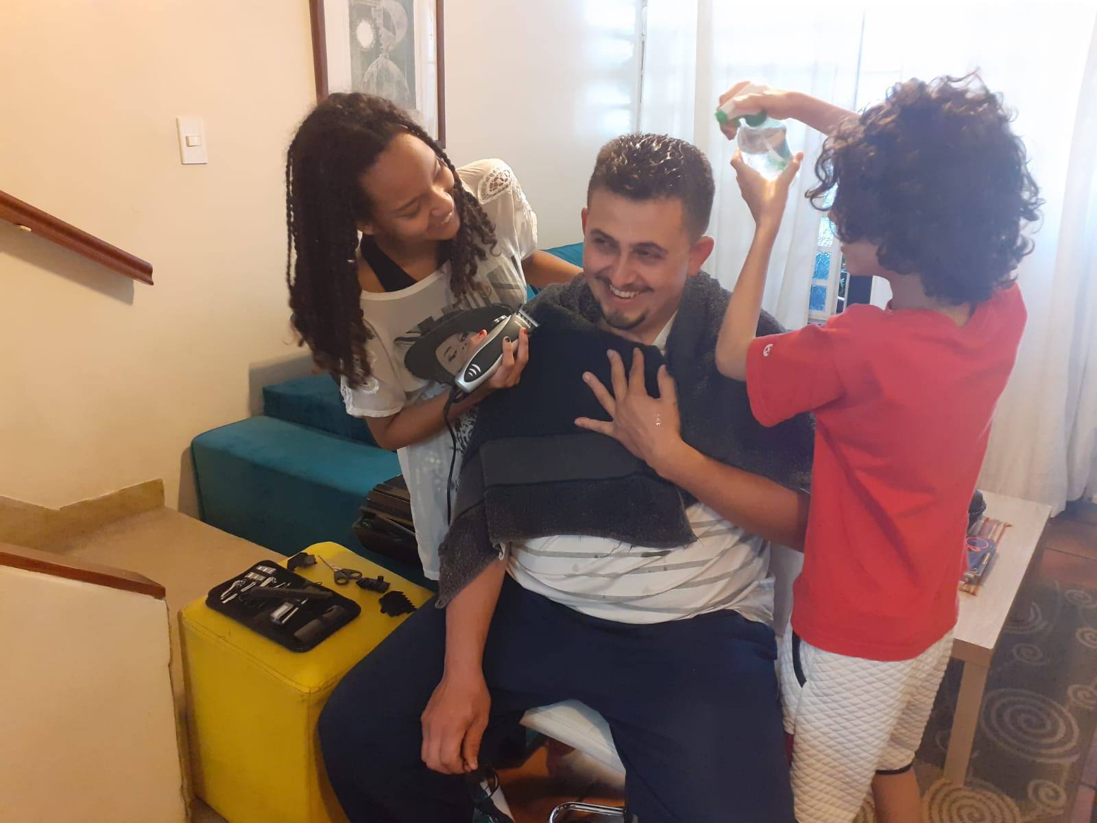 A filha Vitória segura a máquina de raspar o cabelo enquanto o outro filho de Anderson, Ian, borrifa água no cabelo do pai