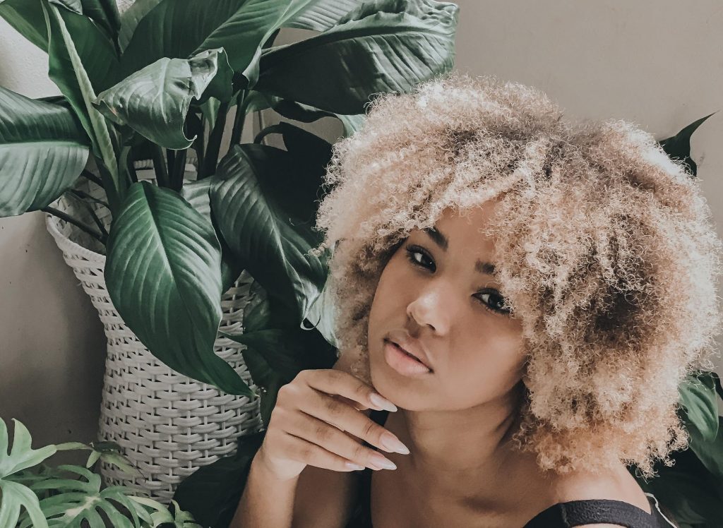 Mulher com cabelo crespo e com tonalidade loiro manteiga, sentada dentro de casa, ao lado de suas plantas