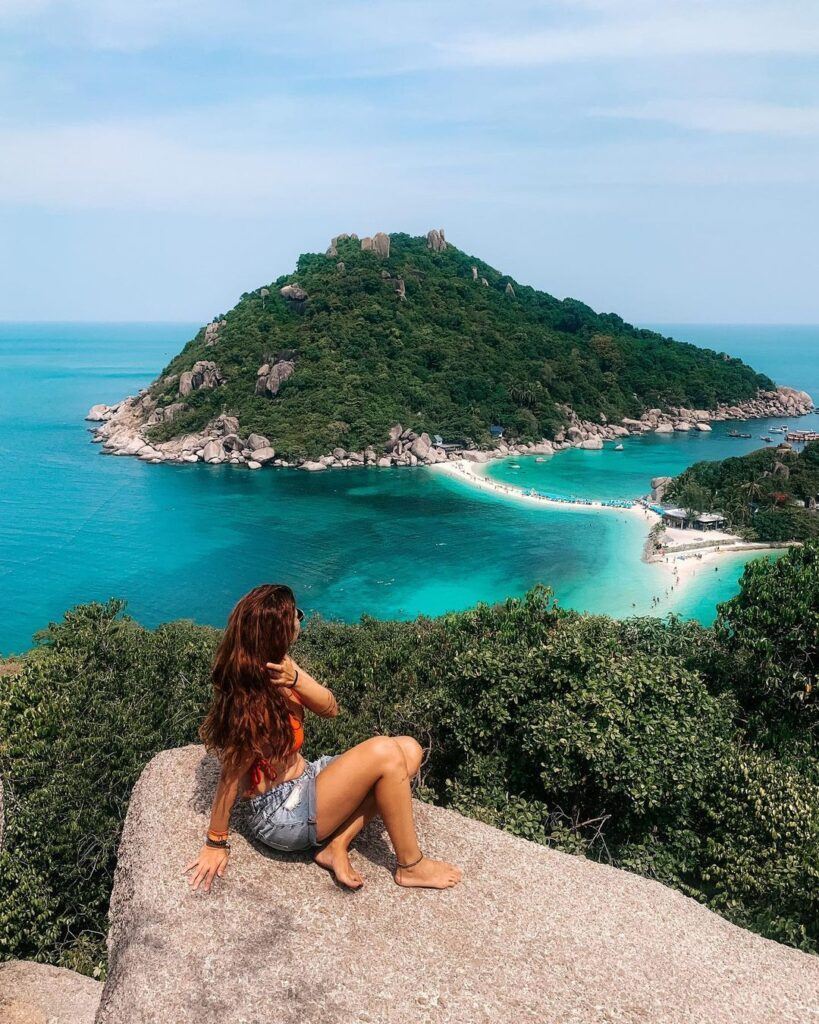 mulher de costas com cabelos soltos sentada em pedra olhando para praia