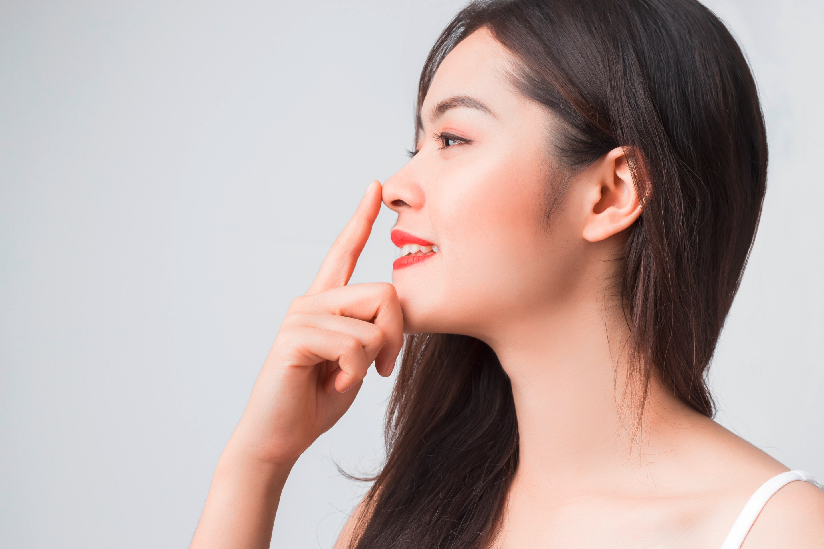 สาวเอเชีย ผิวขาวเหลือง ผมตรง หันข้าง แต่งหน้าโทนสีแดงอมส้ม มือแตะปลายจมูก