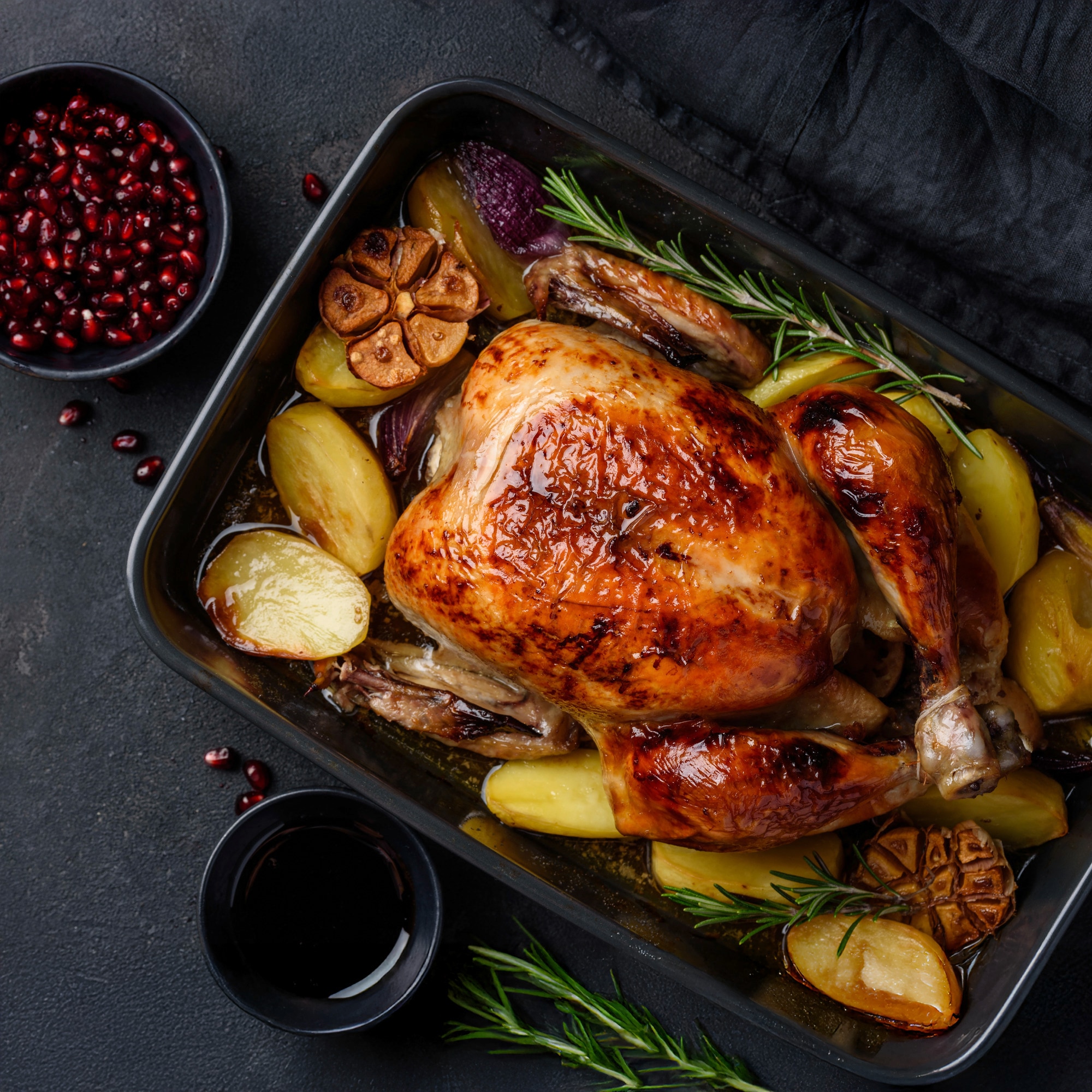 Whole roasted chicken in a roasting dish with potatoes,onions and tall sprig of rosemary with a bowl of pomagranate seeds on the side.