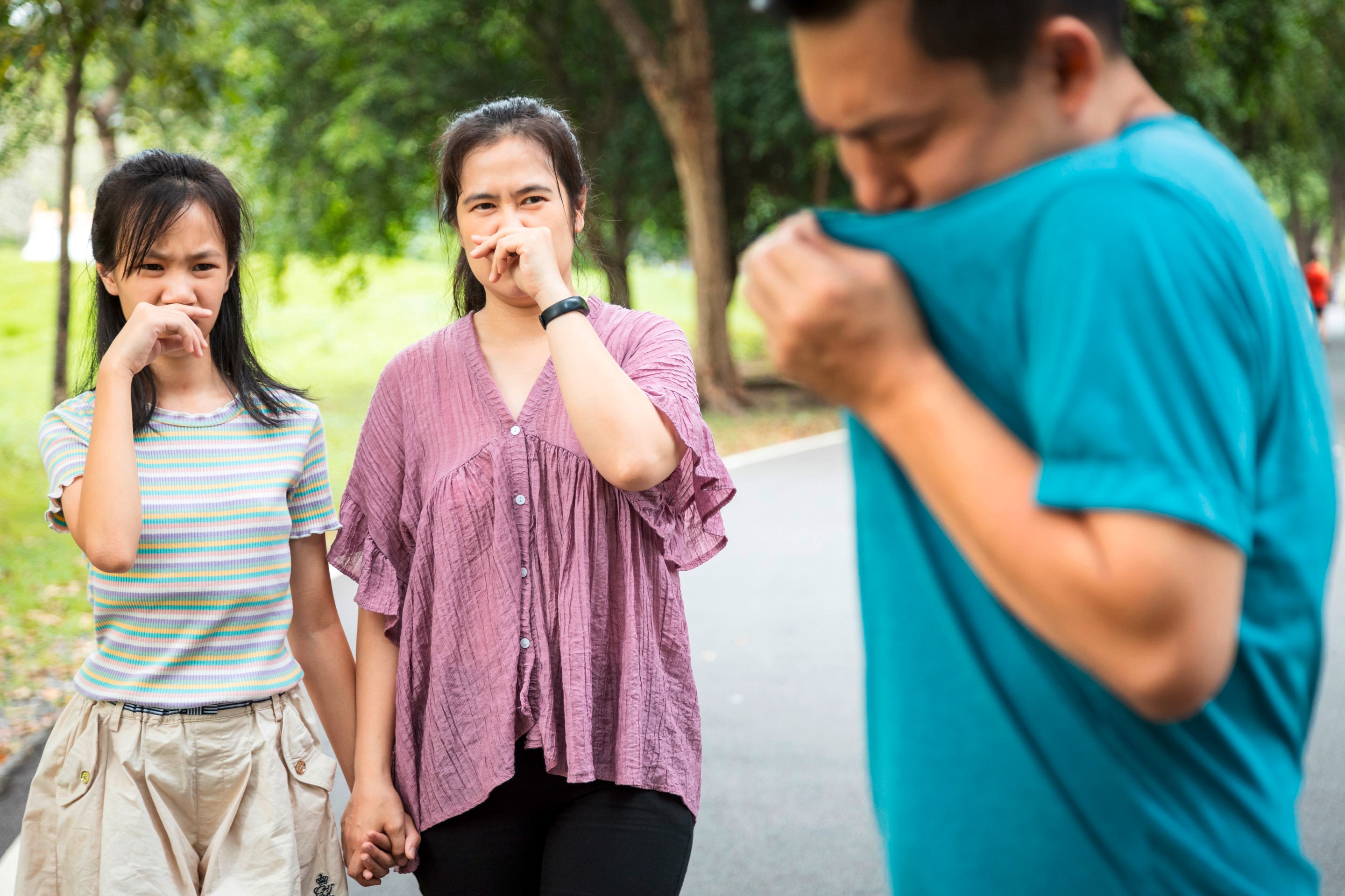 Two women at the park covering noses while looking at man smelling his shirt.