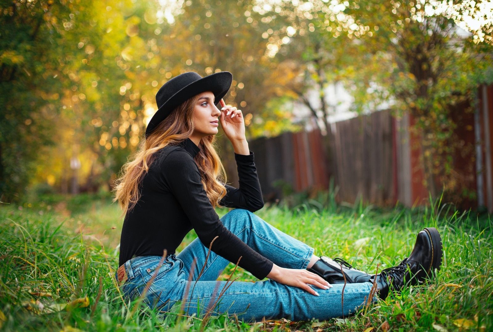 woman with long brown hair, sitting on grass and wearing a big black hat
