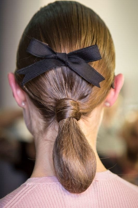 Medium hairstyles: Back view of woman with brown straight hair styled in a ponytail tuck with black bow ribbon.