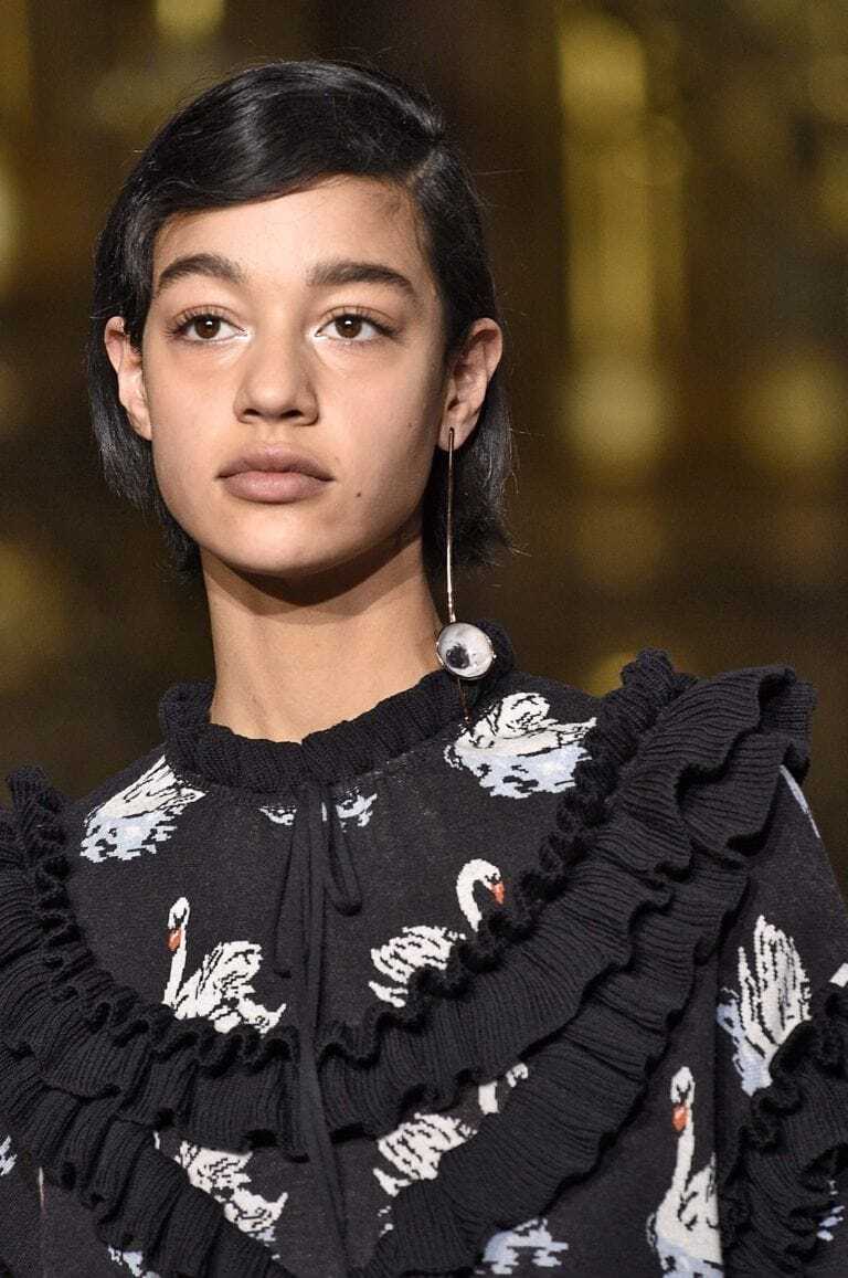 Short hairstyles: Woman on runway with long bob side parted haircut on brown hair. Model is wearing a dark patterned top.