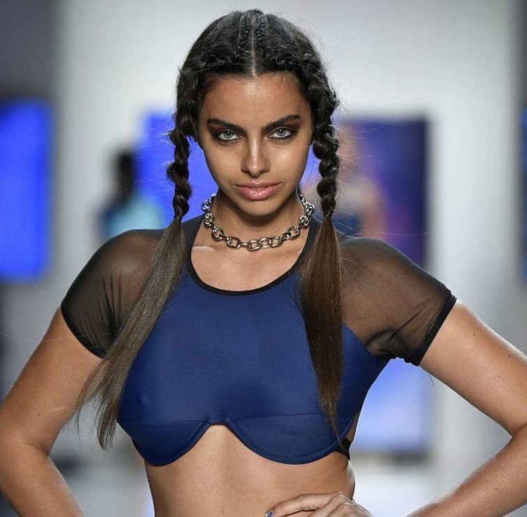 hairstyles for long hair: Woman on runway with long dark brown hair with single corn down the centre and two braided pigtails. Woman is wearing a navy and black mesh sleeve crop top.