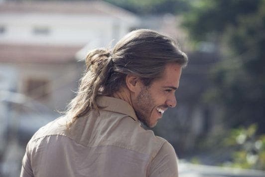 Man bun: Man with long dirty blonde hair in low messy bun wearing a stone coloured shirt.
