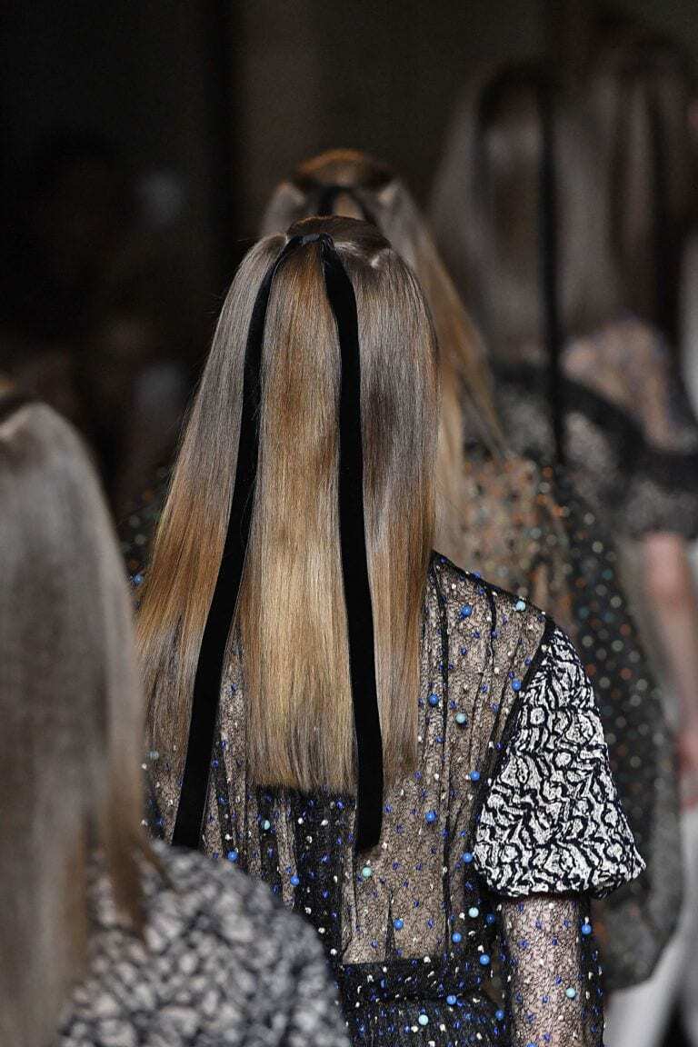 Long hairstyles: Woman with long brown straight hair styled in a half-up, half-down hairstyle with black ribbon. Woman is on the runway wearing a embellished dress.