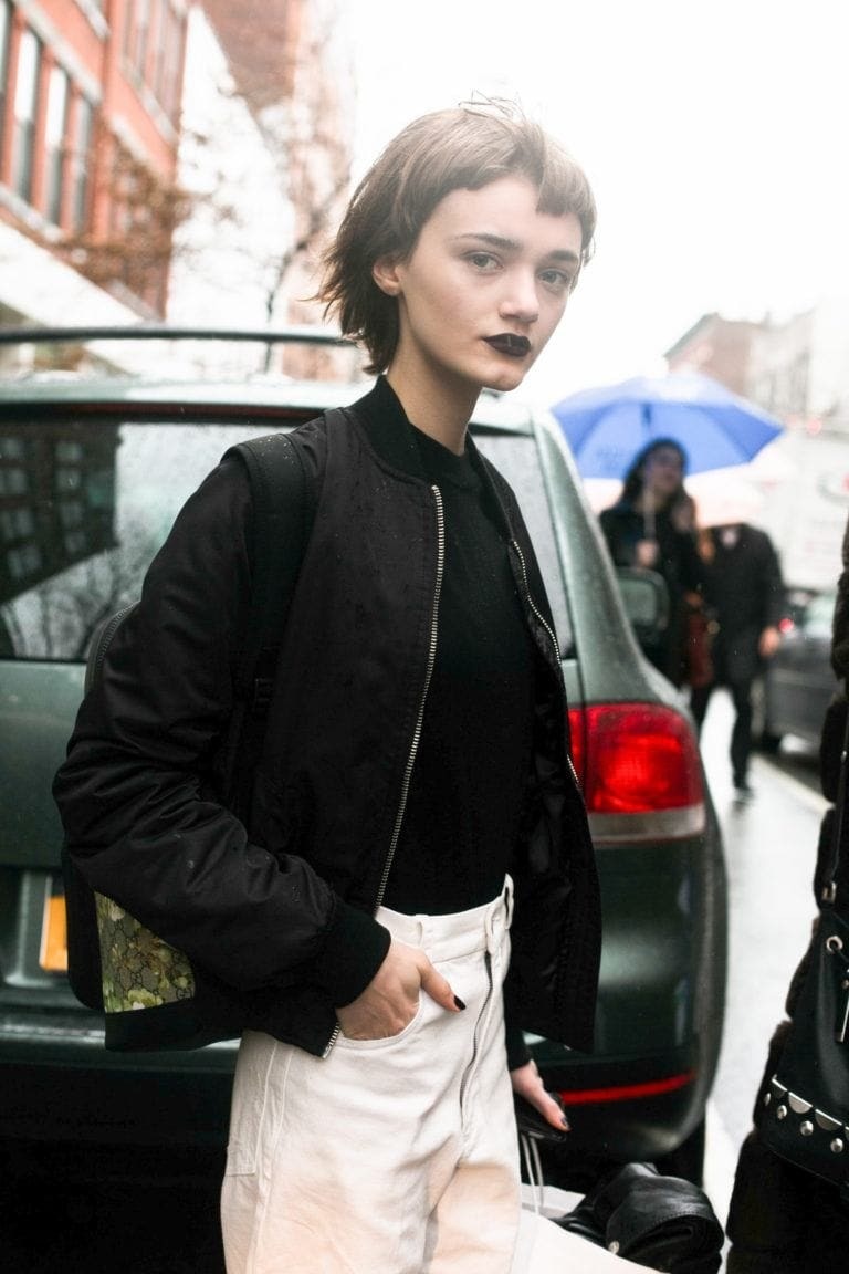 Short hairstyles: Street style image of a woman with short brown hair styled in a bob with short bangs, Woman is wearing a bold dark lipstick and a black baggy jumper and white bottoms.