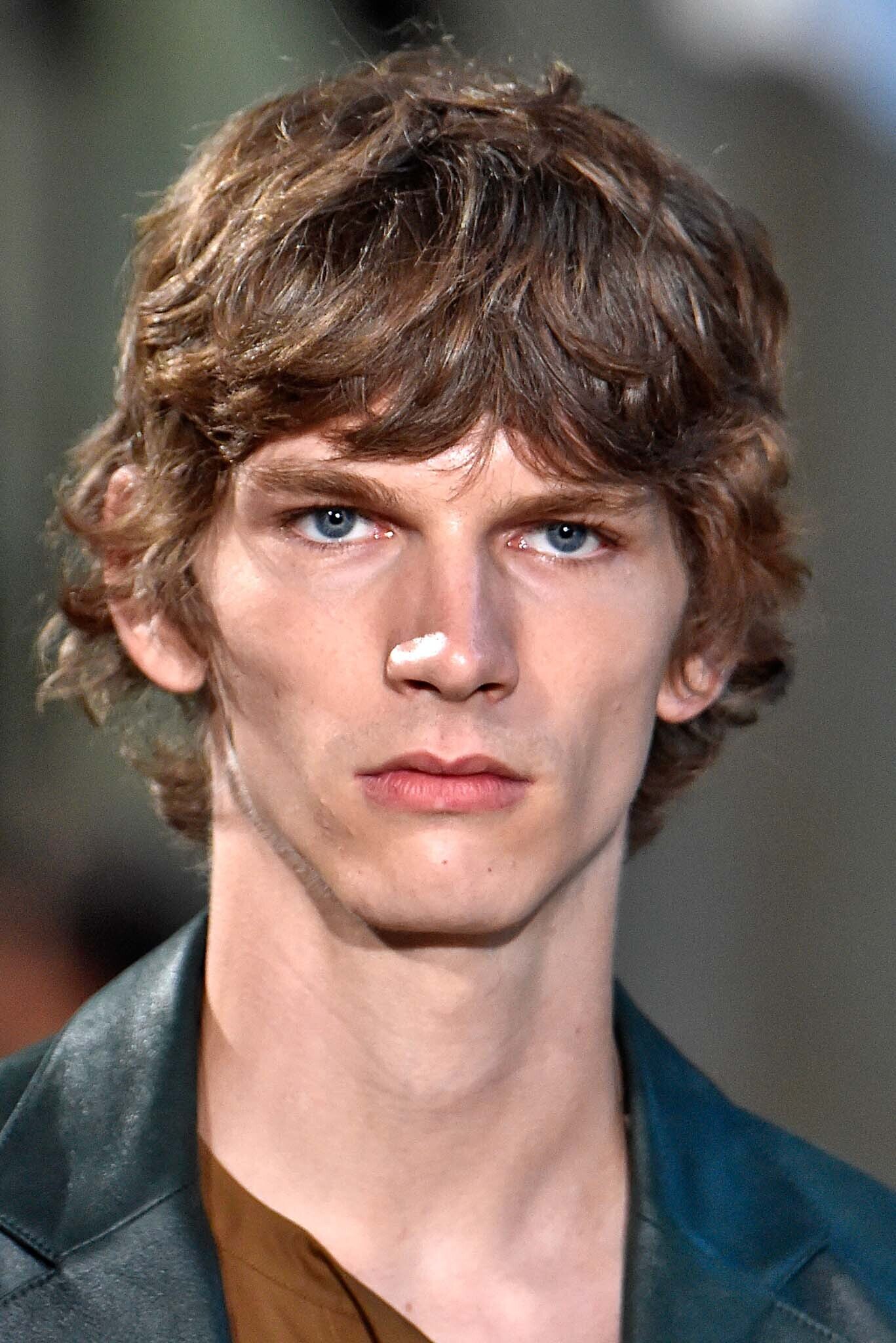 Curly hairstyles for men: Man on runway with brown naturally curly hair with fringe.