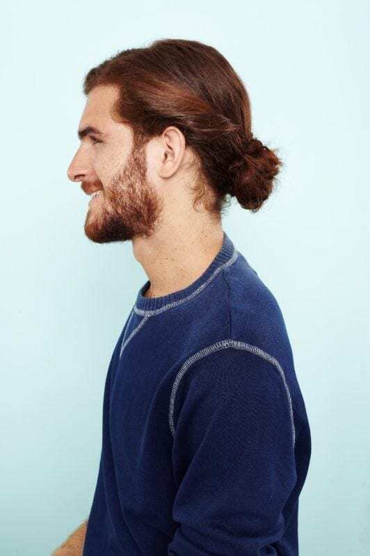 Man bun: Side view of a man with curly auburn hair in twisted low bun wearing a navy blue sweatshirt.