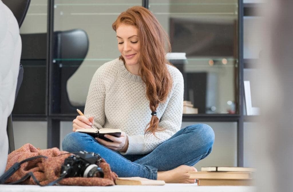 hairstyles for long hair: Woman sitting crossed legged on the floor with long wavy auburn red hair in a side loose braid ponytail while writing surrounded by books.