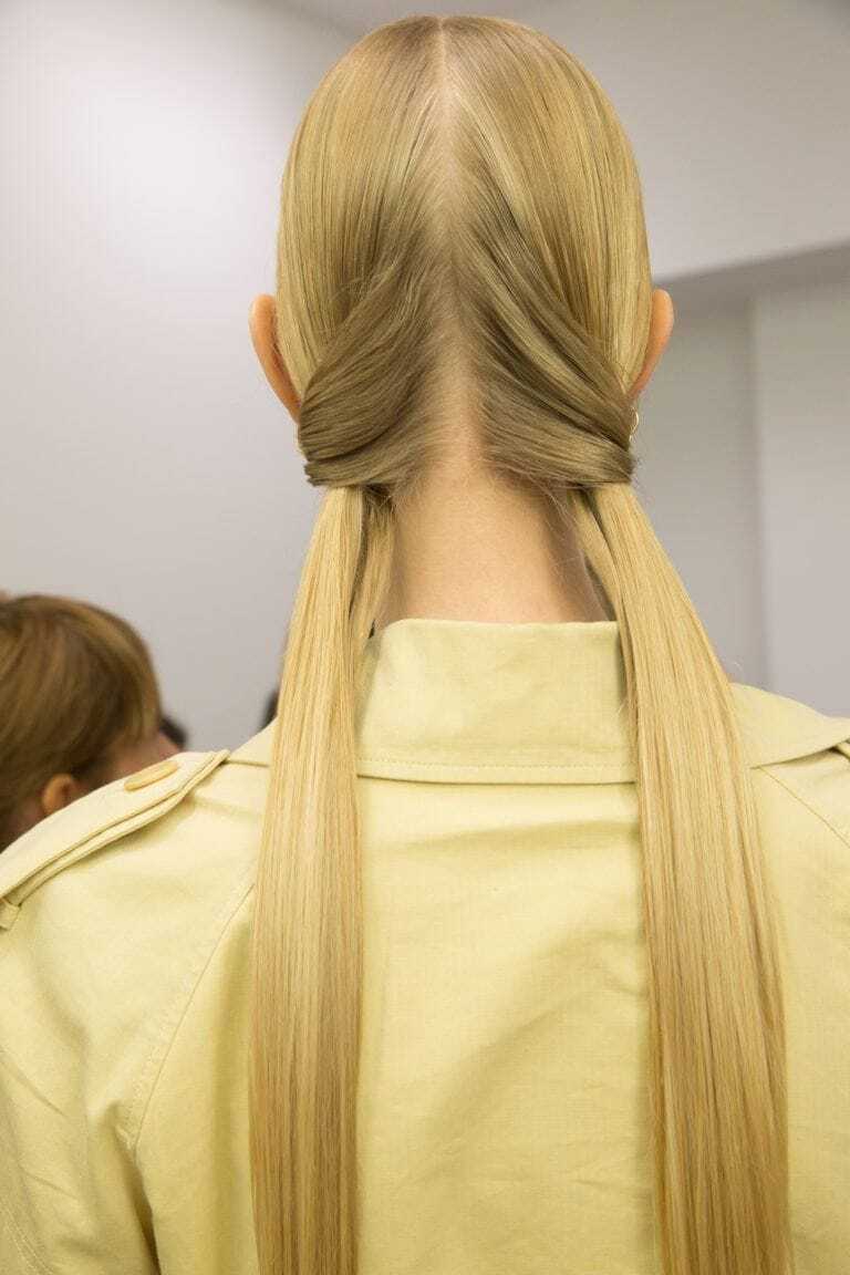 Hairstyles for long hair: Back view of woman with long straight blonde hair in two twisted low pigtails. Woman is wearing a yellow collared jacket.
