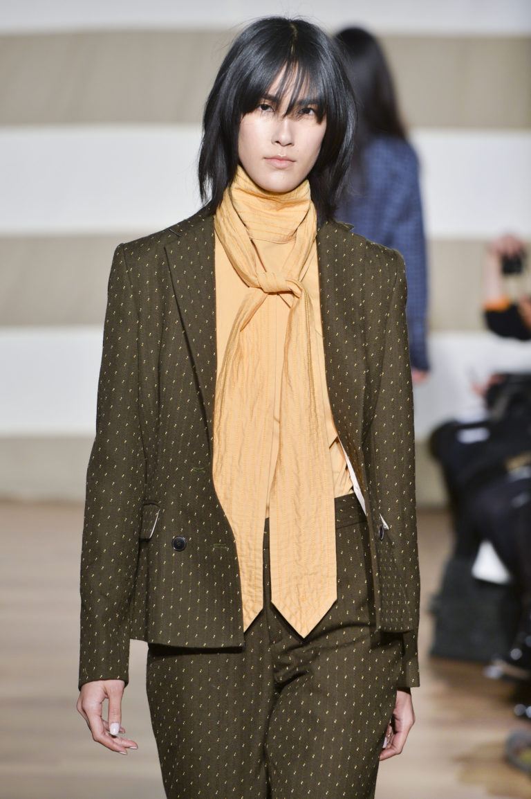 Woman with shoulder length shag haircut on dark brown hair seen in runway image. Woman is wearing a loose fitted suit with a camel coloured scarf.