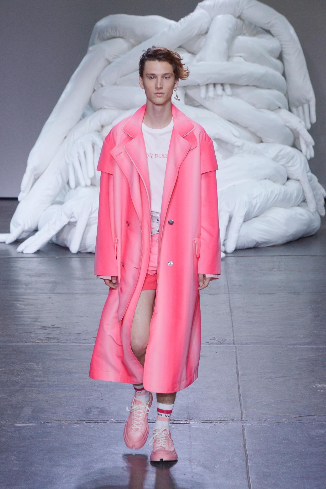 Curly hairstyles for men: Man on runway with brown curly hair swept to one side wearing a long bright pink coat.