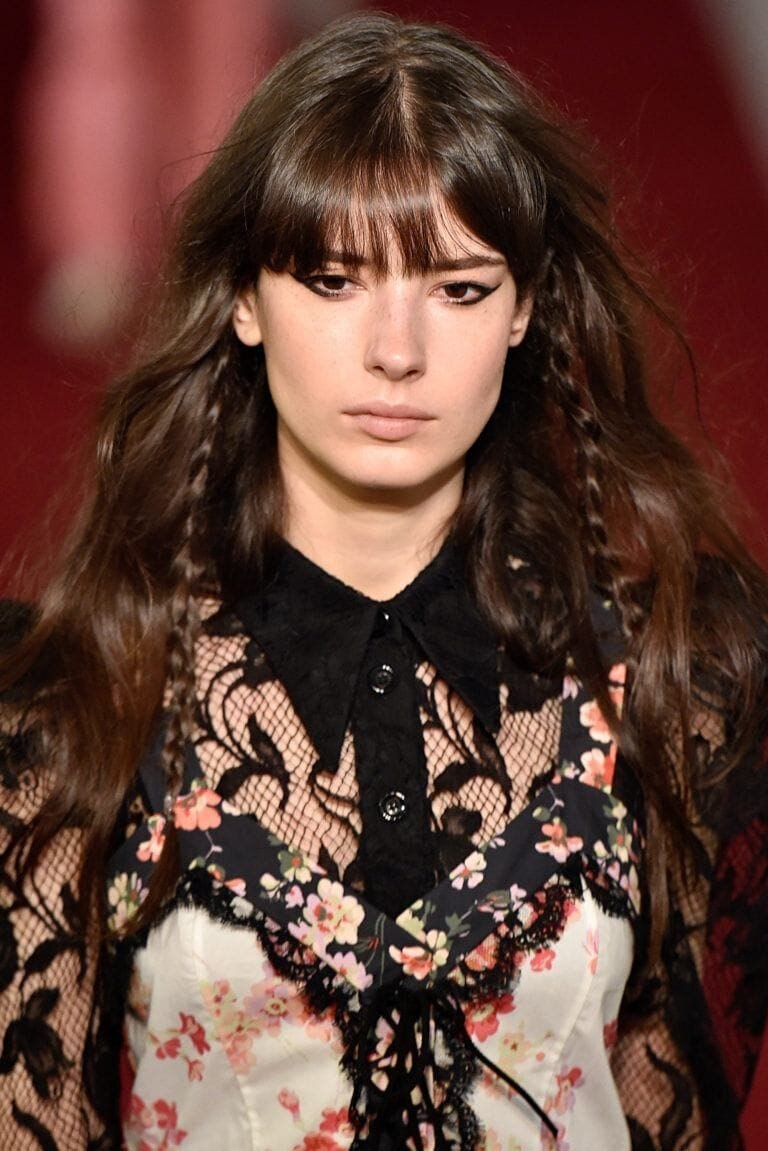 Long hairstyles: Woman on runway with long wavy brown hair with long bangs and peekaboo braids. Woman is wearing a floral and lace top.
