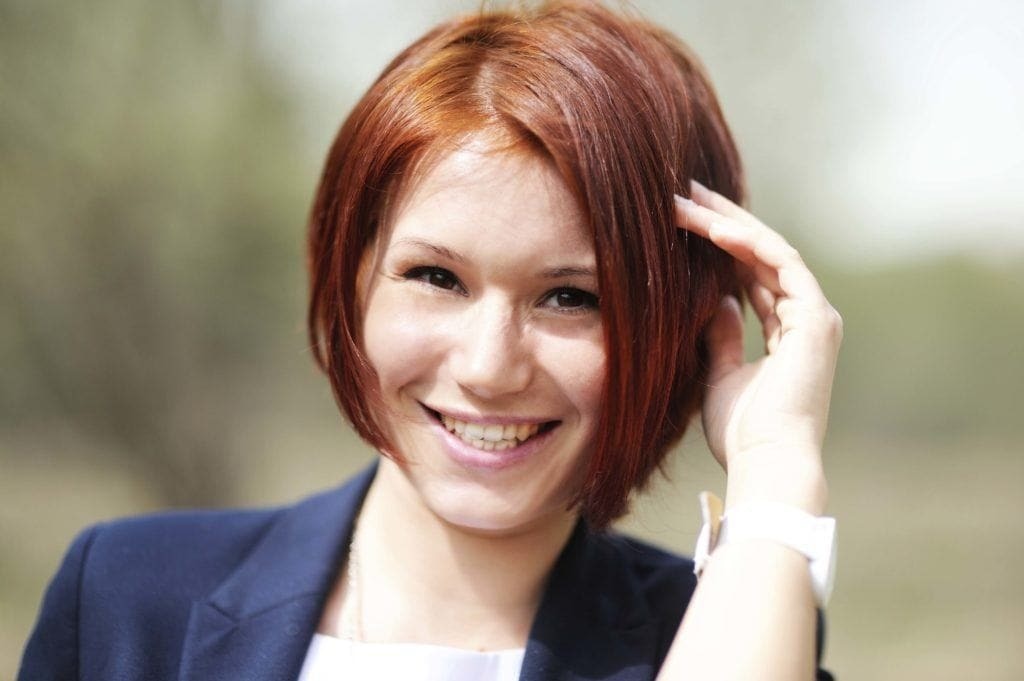 Woman with auburn red straight hair styled in a side parted chin grazing bob. Woman is touching her hair and wearing a navy blazer jacket.