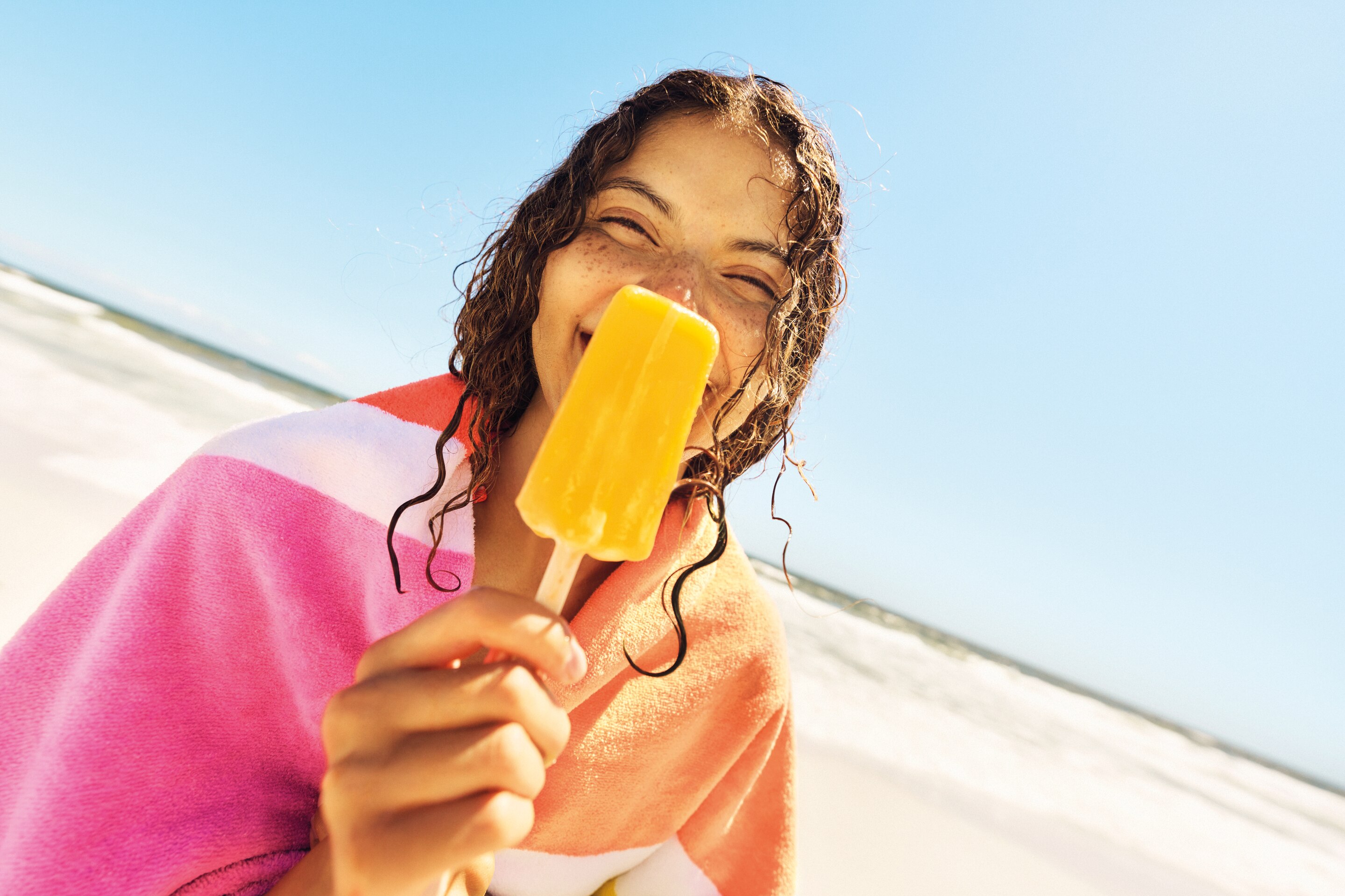 Person at the beach enjoying a bright yellow Magnum popsicle against a clear blue sky