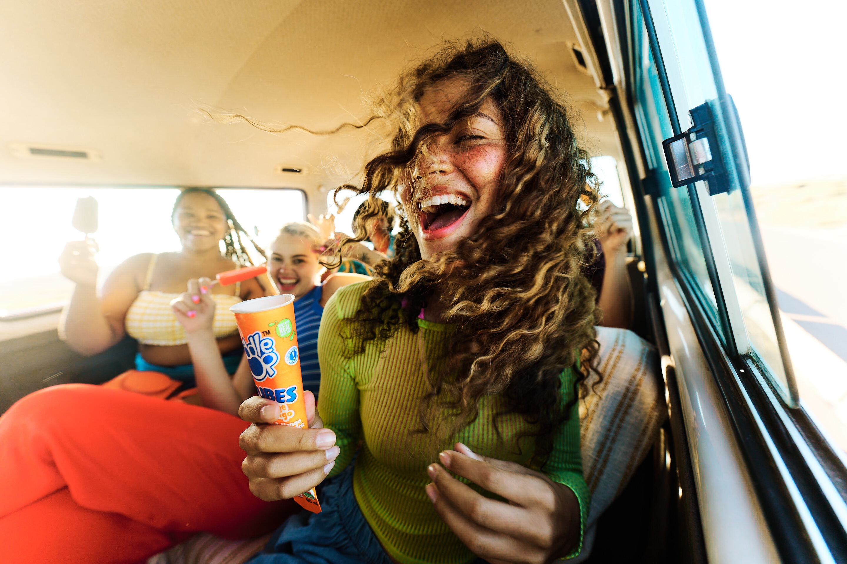 Laughing person in green top holding a popsicle in a car with friends in background.