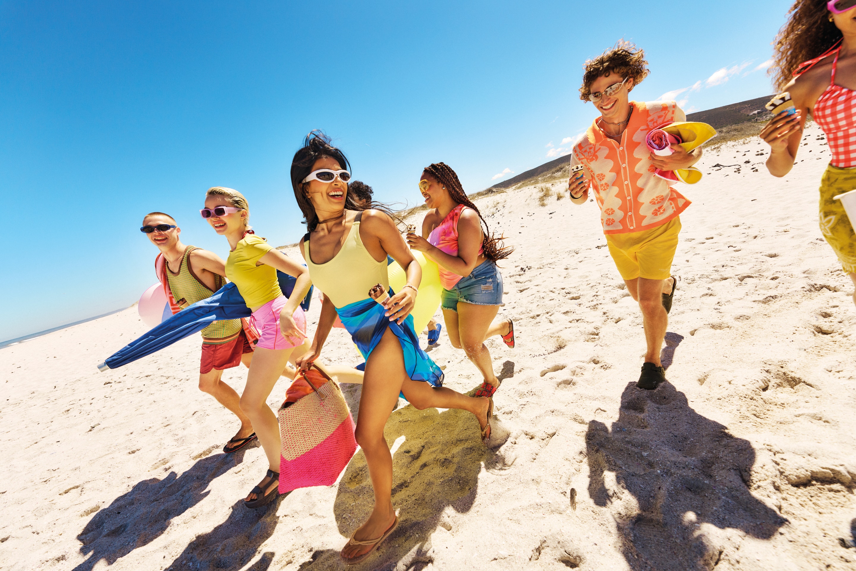 Group of friends running on beach with colorful swimwear and holding a Cornetto on sunny day.