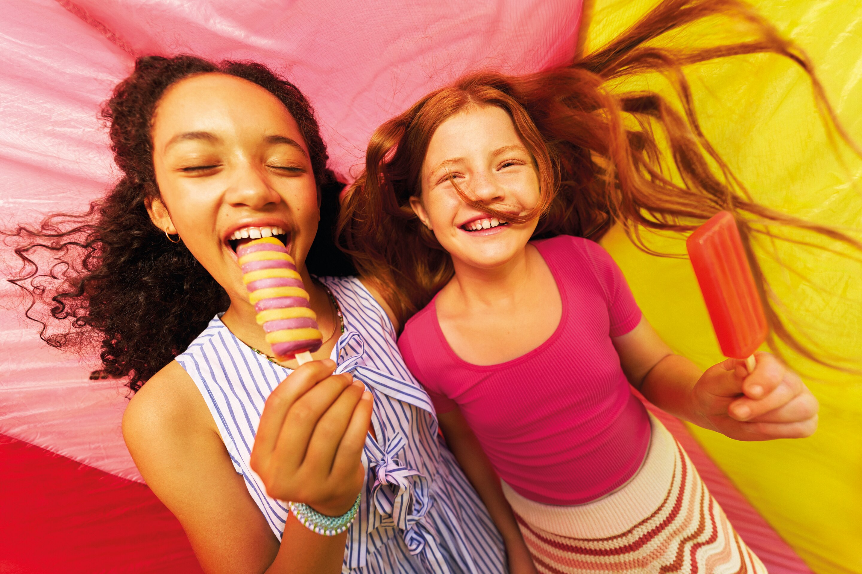 Two smiling children with curly hair holding colorful Twister ice cream against a pink background.