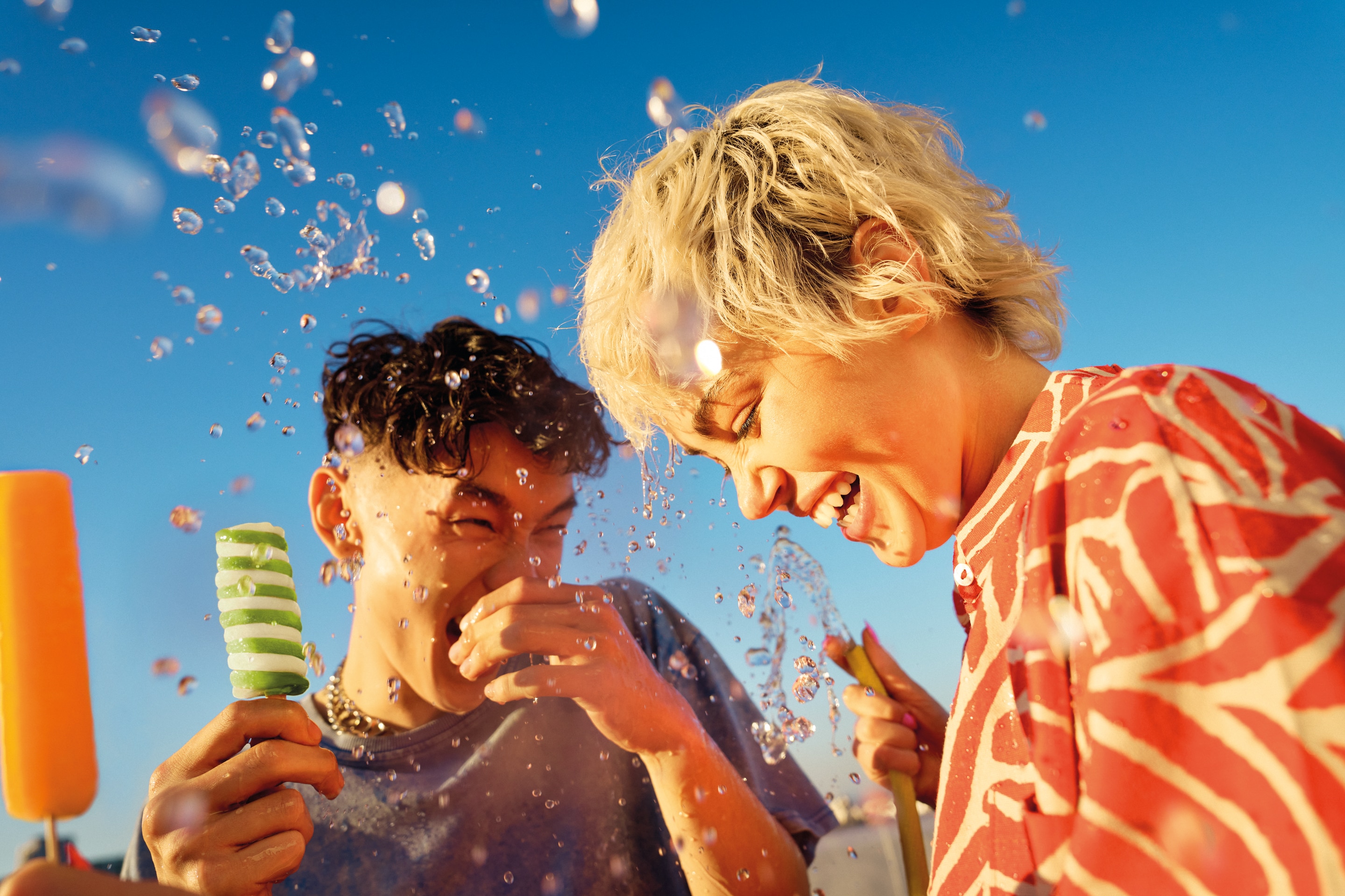 Two young people halding Twister ice creams amid water splashes against bright blue sky.