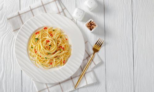 Resep aglio olio disajikan dalam piring putih di atas serbet dan meja kayu putih.