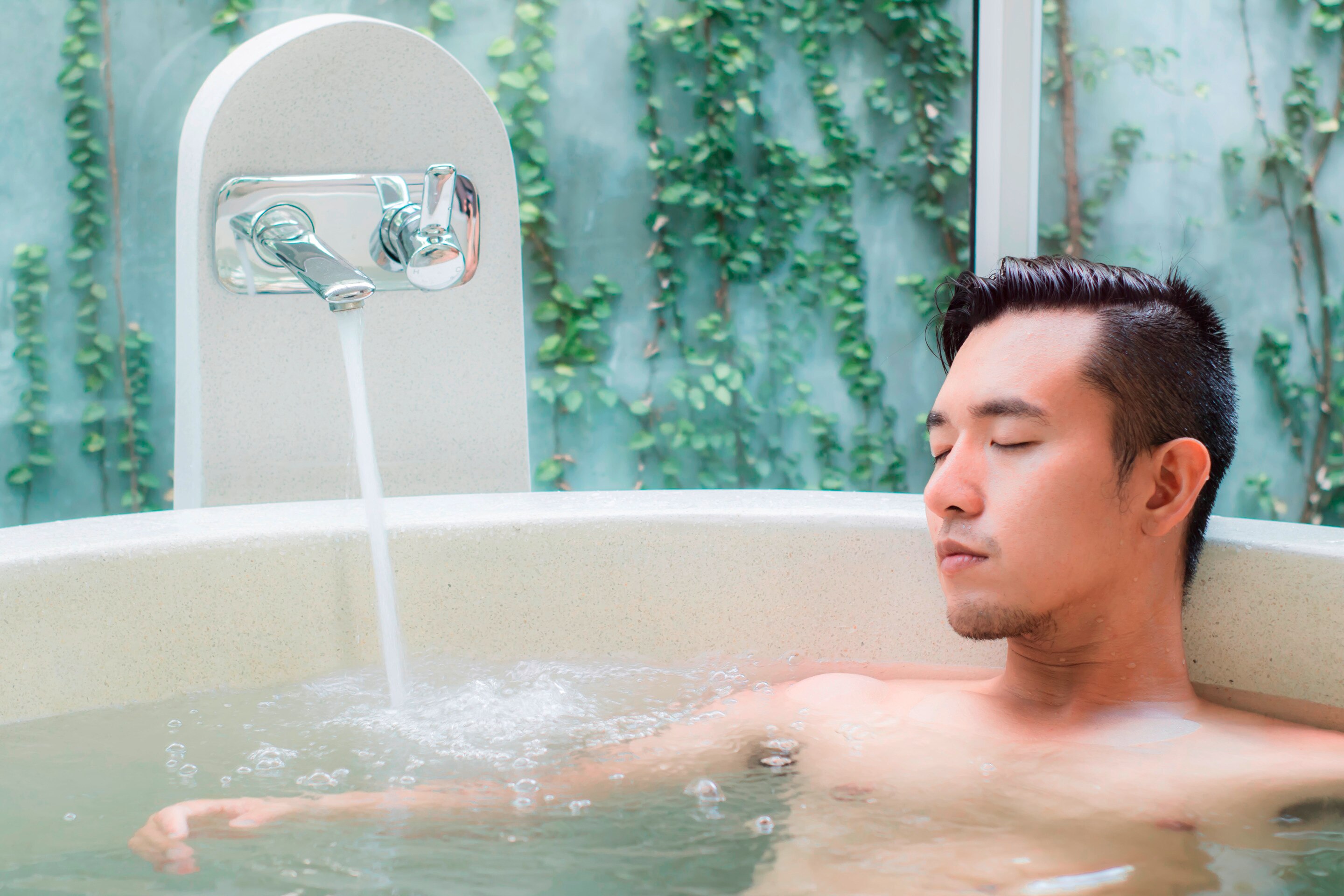 Asian man relaxing in tub with faucet on.