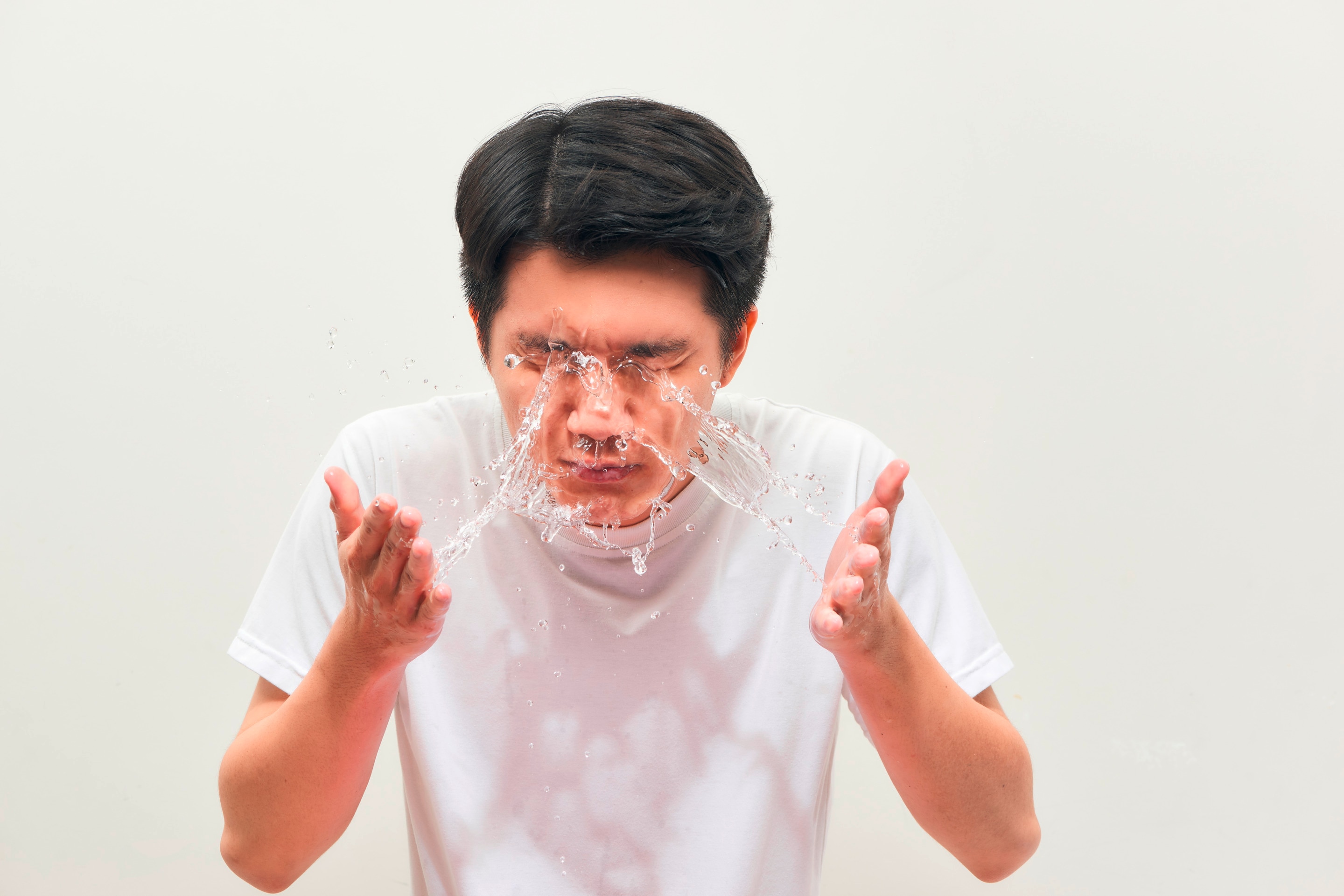 Asian man in white shirt splashing water on face.
