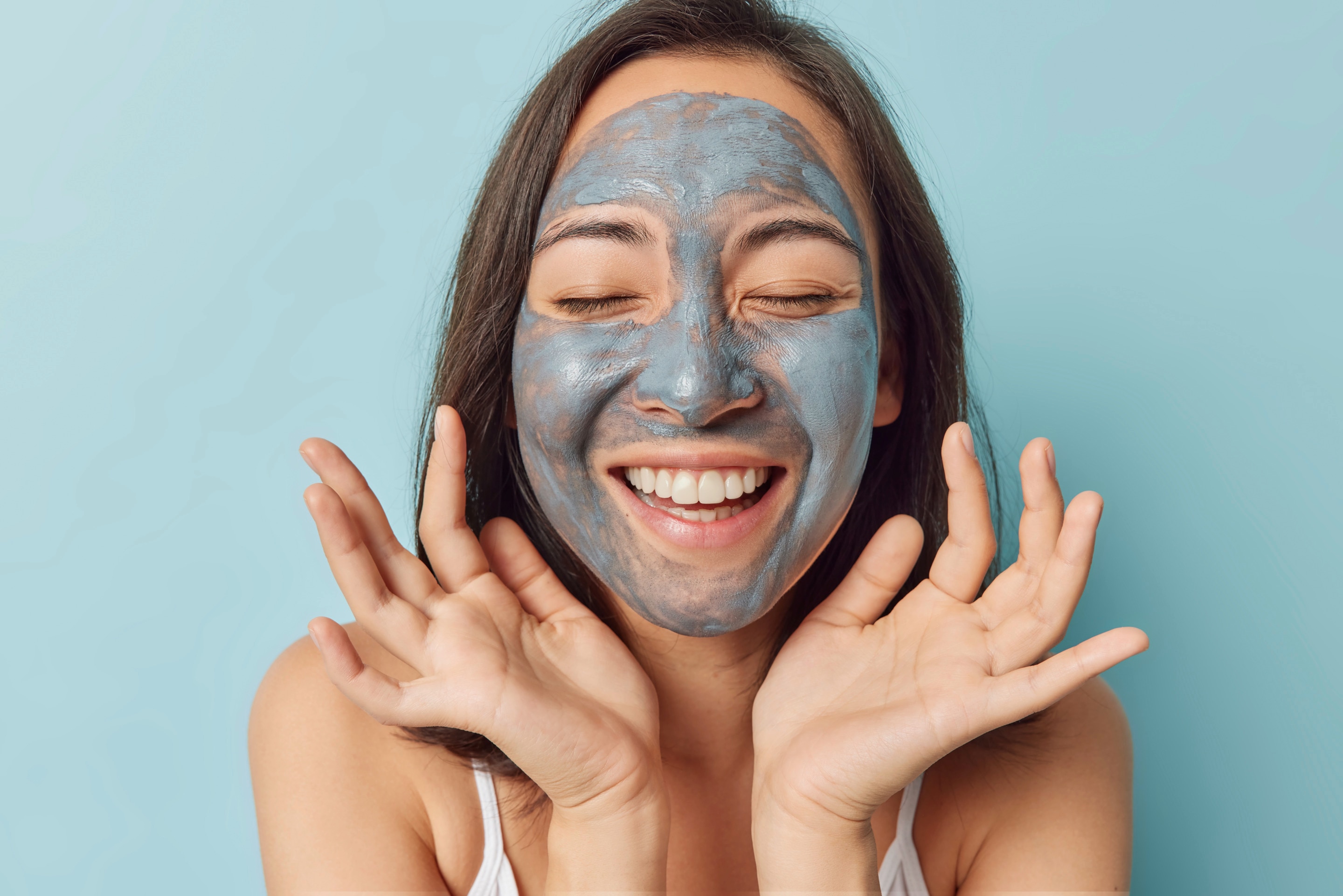 ผู้หญิงเอเชีย ผมประบ่า แสกข้าง ผมสีน้ำตาล หน้าตรง หลับตา ยิ้มตาหยี ยิ้มเห็นฟัน บนหน้าทามาส์มากสีฟ้าอมเทา สองมือหงายรับกับช่วงใบหน้าแบบน่ารัก