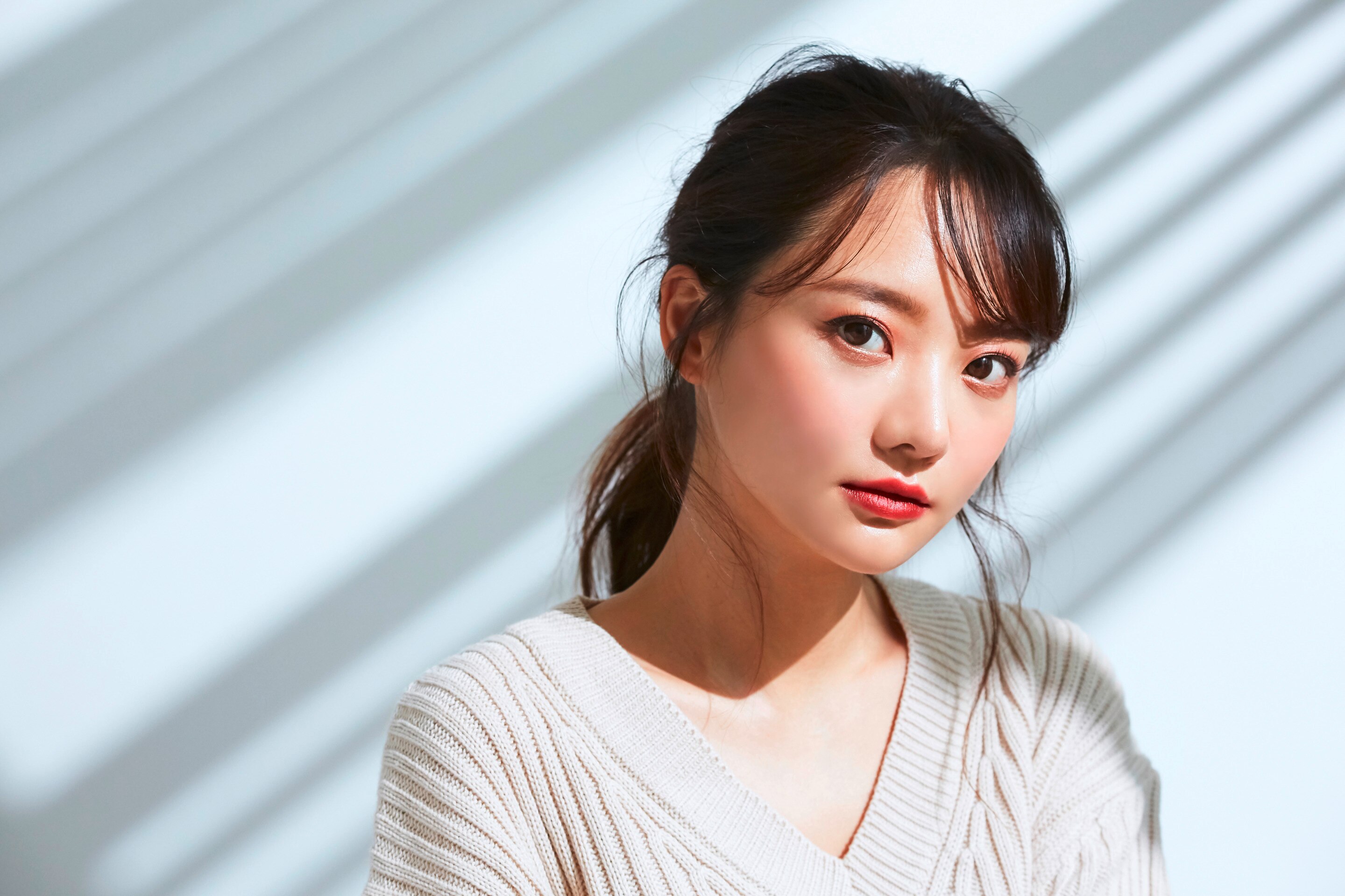 ผู้หญิงเอเชีย หน้าเอียง ผมหน้าม้าปัดข้าง รวบผม มีปอยผมด้านข้าง ทาลิปสีแดงอมชมพู สวมสเว็ตเตอร์คอวีสีขาว ด้านหลังมีเส้นเงาพาดเฉียง